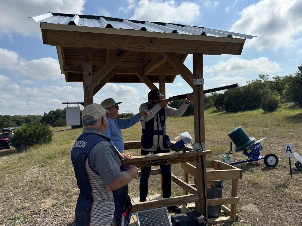 A coach pointing out a target to a shooter mounting his shotgun at a sporting clays stand